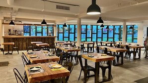 A spacious dining area with multiple tables and chairs, large windows offering views of the outdoors, and a bar area with a counter and coffee machines.