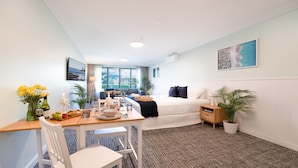A hotel room with a bed, a desk with a chair, a dining table with chairs, a small table with a vase of flowers, and a large window with a view of greenery.