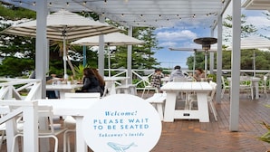An outdoor seating area with white tables and umbrellas, a sign indicating a takeaway service, and a view of the sky and trees.