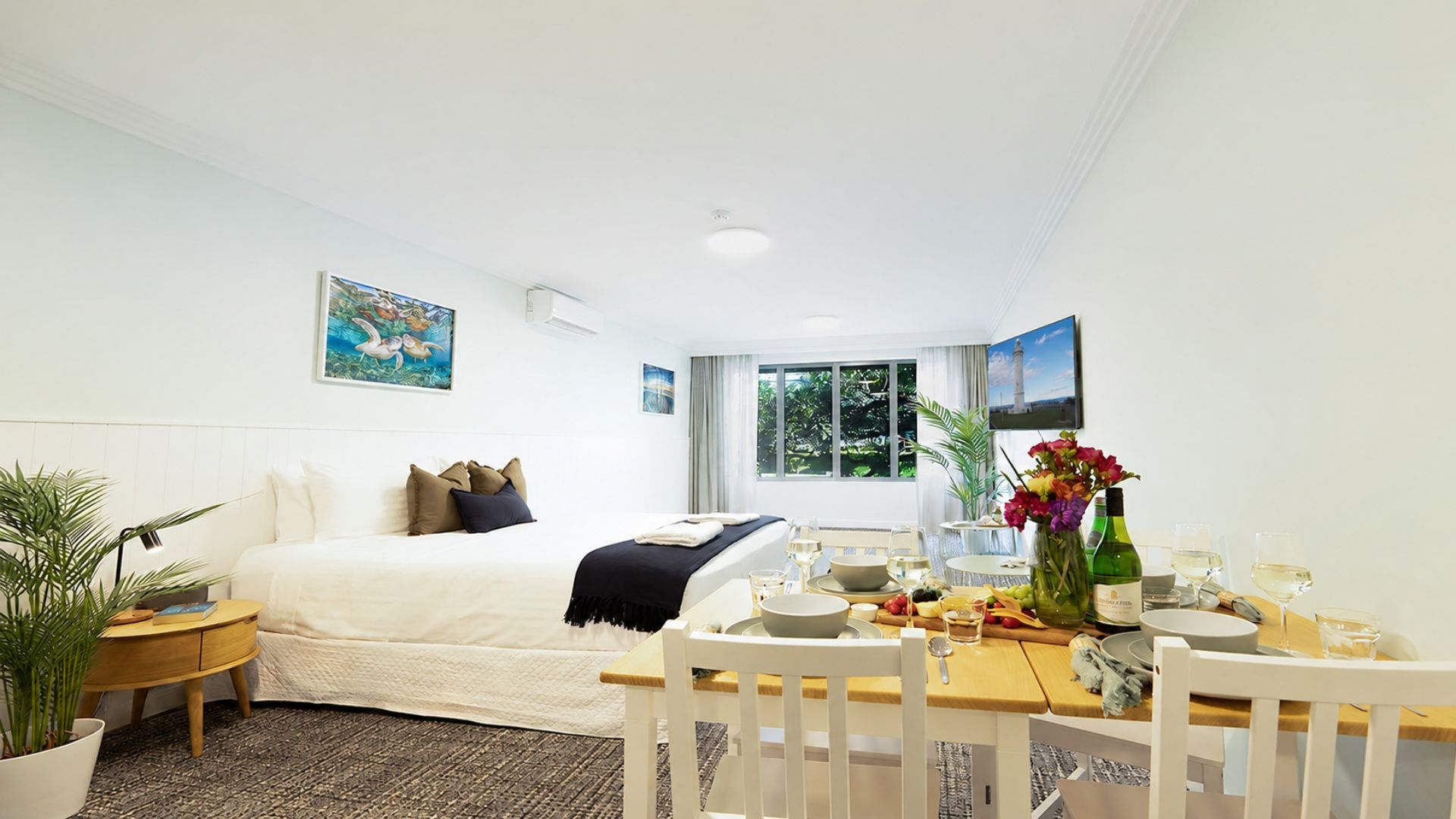 A hotel room with a bed, a dining table with chairs, a potted plant, and artwork on the walls.