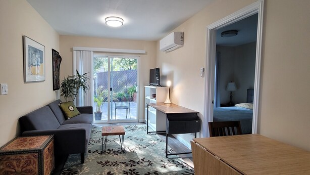 A compact living space with a sofa, a small table, a desk, and a TV. There is a sliding glass door leading to an outdoor area with plants. - Charming 1-bedroom cottage with AC near Piedmont and Lake Merritt. Pet friendly! (Oakland)
