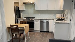 A compact kitchen with white cabinets, a stove, and a dishwasher. There is a dining table with chairs and a gray sofa. - Charming 1-bedroom cottage with AC near Piedmont and Lake Merritt. Pet friendly! (Oakland)
