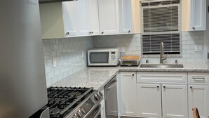 A modern kitchen with white cabinets, stainless steel appliances, a microwave, and a window with blinds. - Charming 1-bedroom cottage with AC near Piedmont and Lake Merritt. Pet friendly! (Oakland)