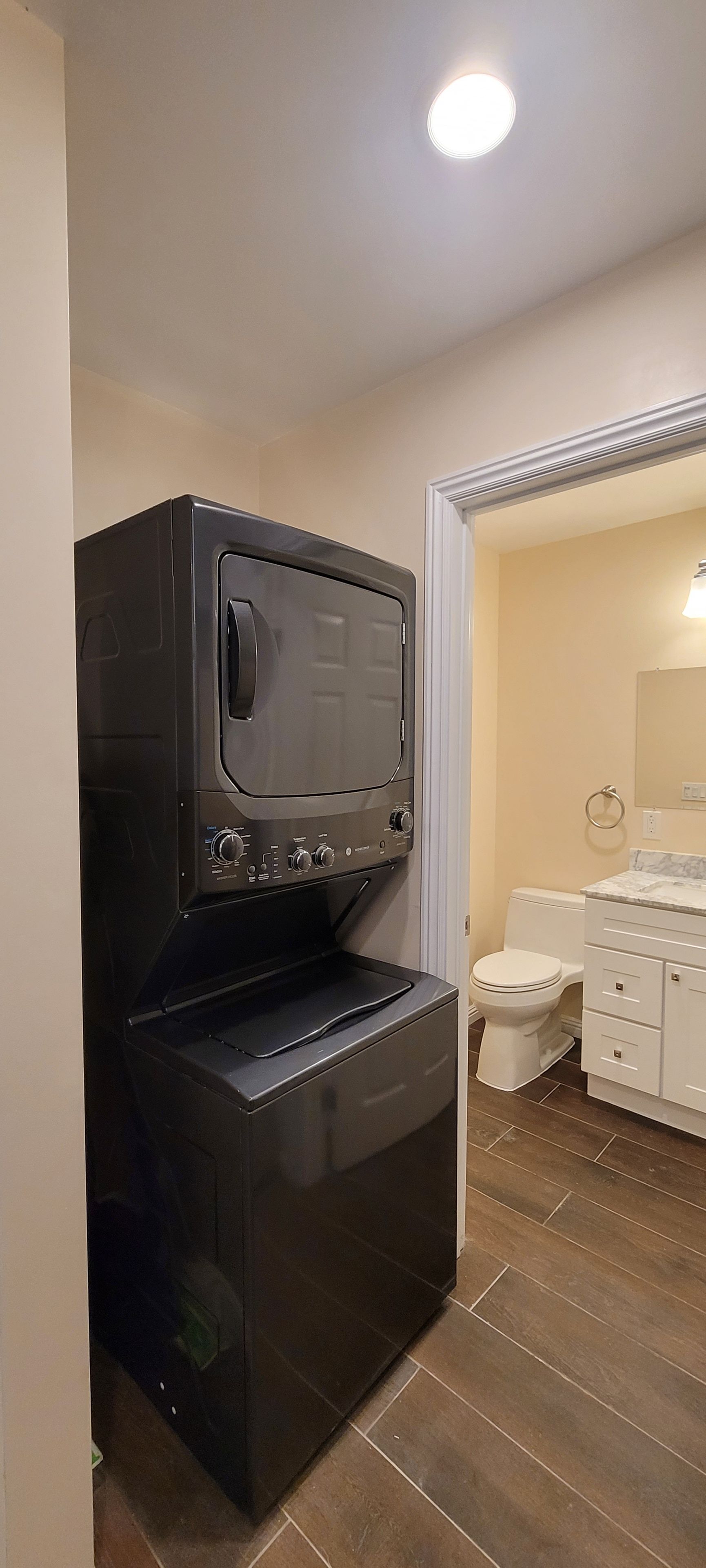 Un lave-linge et un sèche-linge compacts, de couleur foncée, sont placés dans un coin d’une pièce dotée d’un plancher en bois et d’une porte blanche donnant sur une autre pièce.