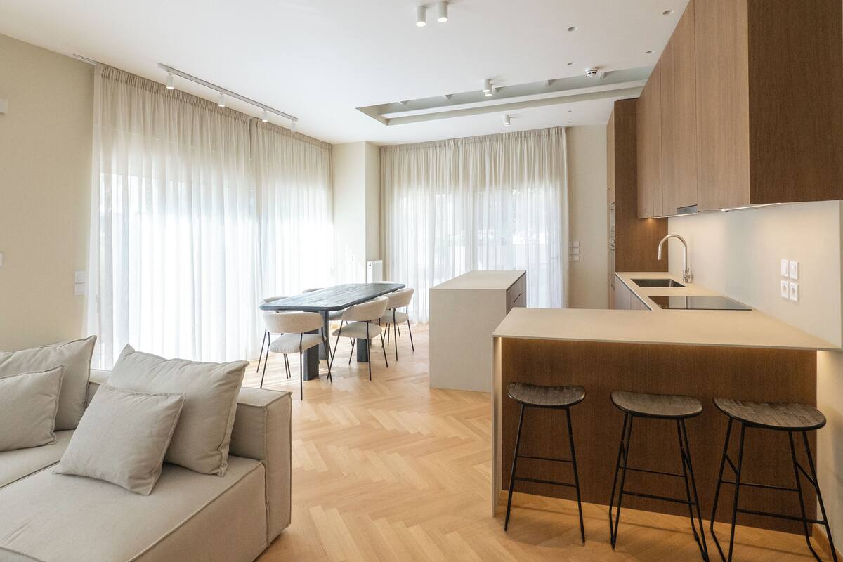 A modern kitchen with a central island, bar stools, a dining area with a table and chairs, and a sofa in a living area.