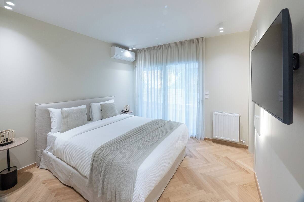 A modern bedroom with a large bed, a bedside table, a wall-mounted TV, and a window with curtains.