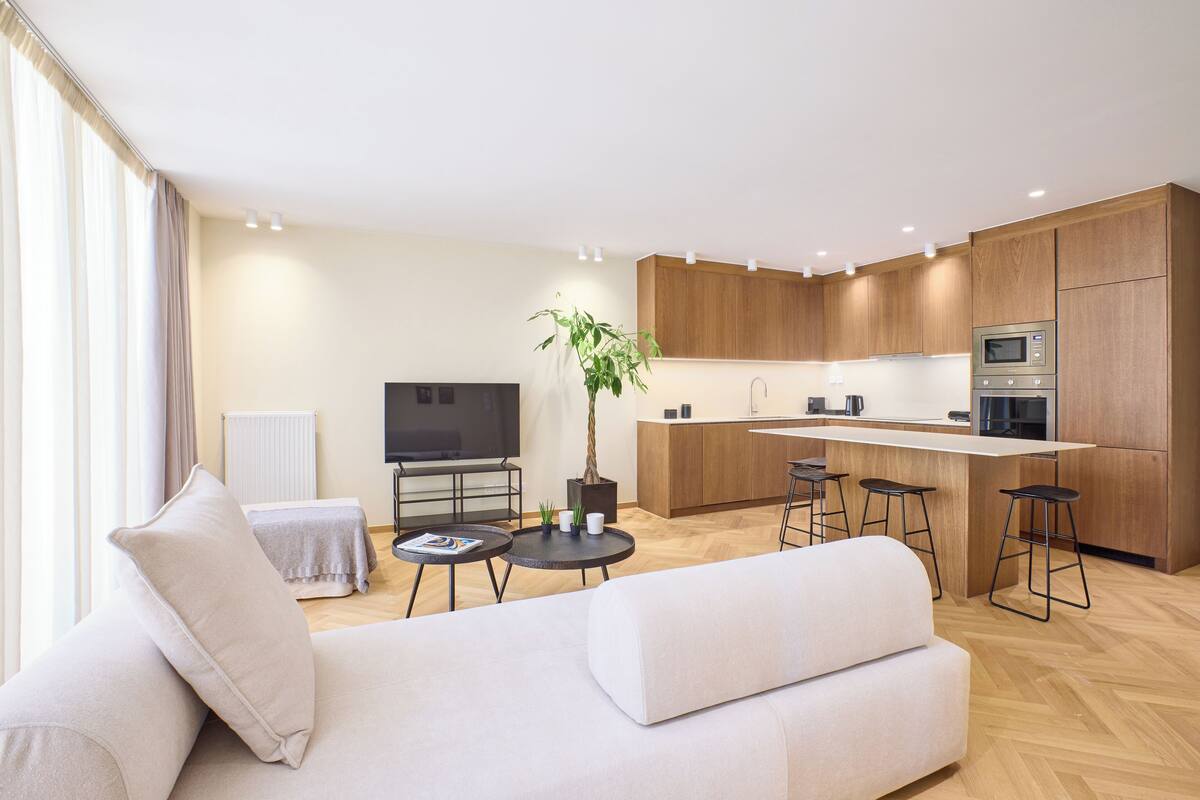 A modern living room with a sofa, a coffee table, and a TV. There is a kitchen area with wooden cabinets and a microwave.