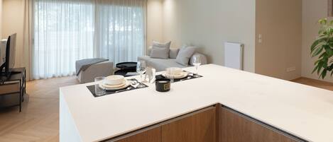 A modern kitchen with a central island, a dining area with a sofa, and a large window with sheer curtains.