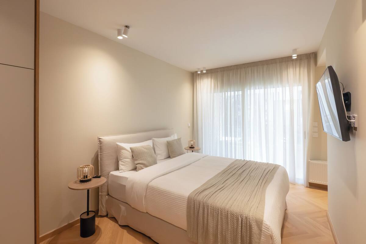 A modern bedroom with a large bed, a bedside table, a TV mounted on the wall, and a window with sheer curtains.