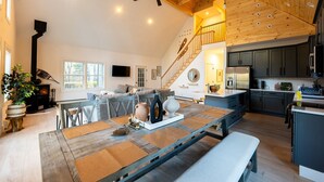 A spacious living area with a high ceiling, a fireplace, a dining table with placemats, a kitchen with dark cabinets, and a staircase leading to an upper level. - The Perfect Modern Killington Getaway
Hot tub-Sauna-Movie Room-Speakeasy-VIEWS! (Killington)
