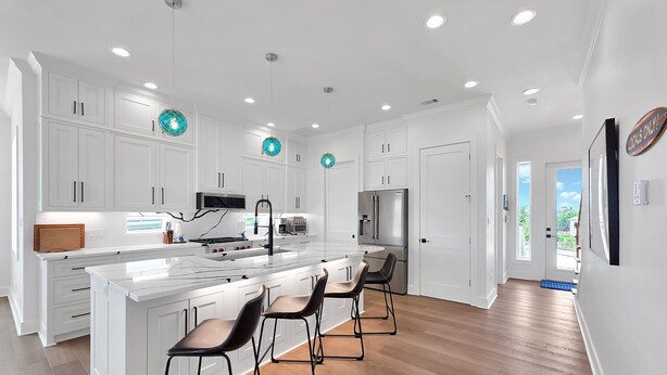 Una cocina moderna con muebles blancos, una isla central con taburetes y electrodomésticos de acero inoxidable, además de iluminación colgante.