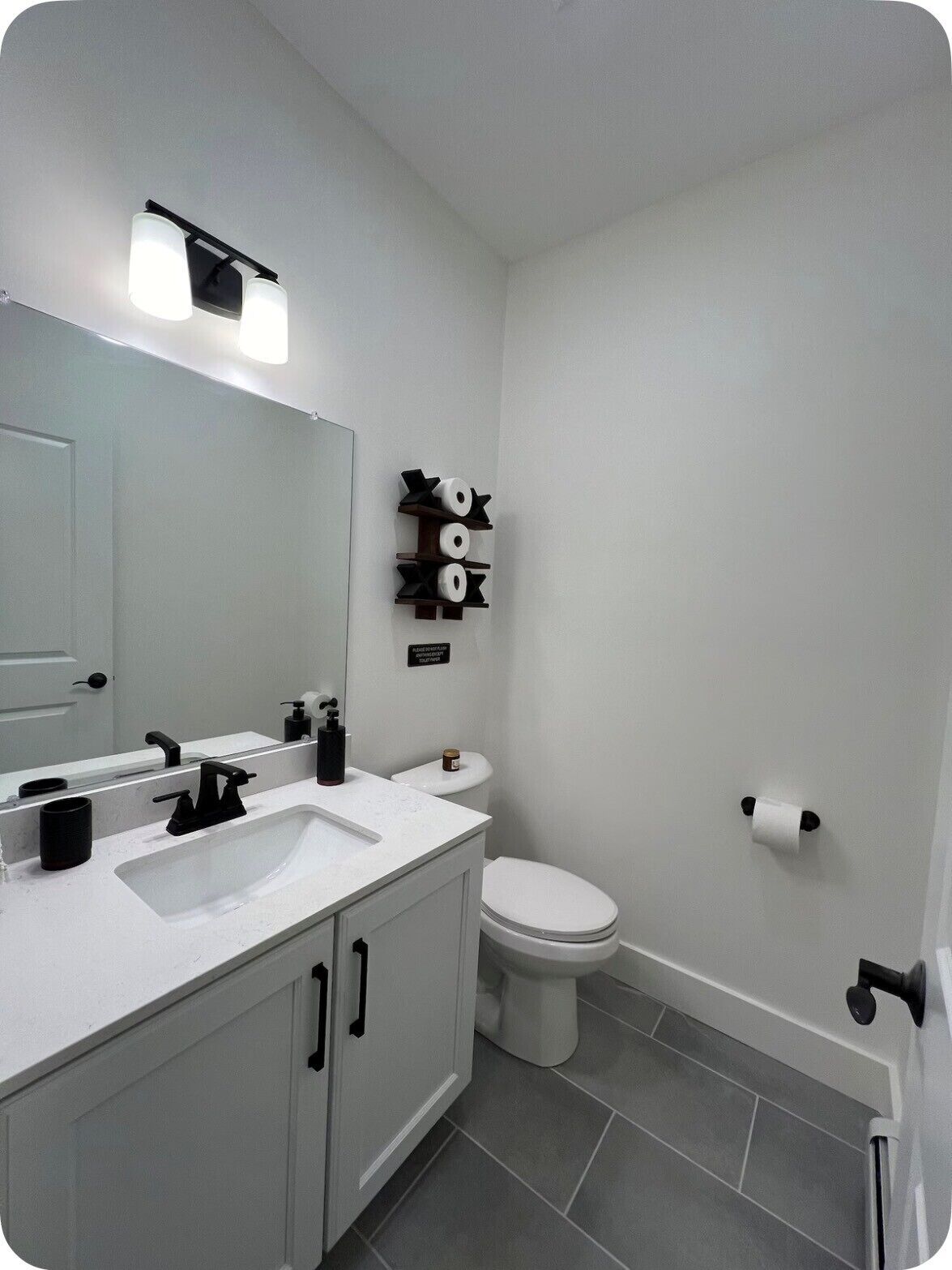 A modern bathroom with a white sink, black faucet, and a toilet. There is a mirror above the sink and a shelf with toilet paper and a sign above the toilet.