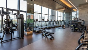 Ein modernes Fitnessstudio mit Laufbändern, Kraftgeräten und einer Bank. Große Fenster bieten Blick auf die Stadt.