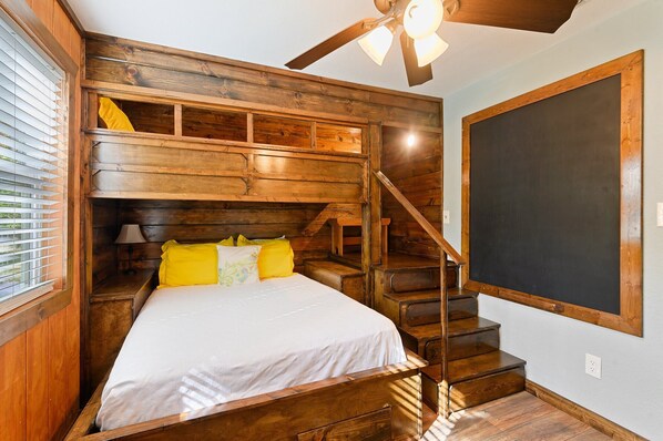 Une chambre de style cabane en bois, comprenant un lit, une table de chevet, une lampe, une fenêtre avec stores, un escalier en bois et un ventilateur de plafond.