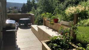 Een houten terras met een barbecue, een tafel en stoelen. Verhoogde bloembedden met verschillende planten en bloemen.