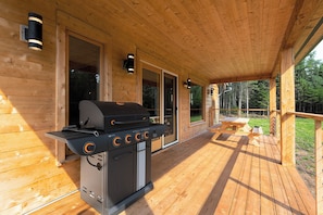 A covered wooden deck with a grill, outdoor seating, and a view of the forest.