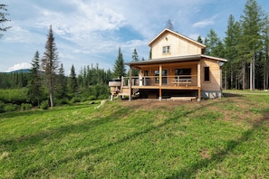A wooden cabin with a balcony, surrounded by a forest and a grassy hill.