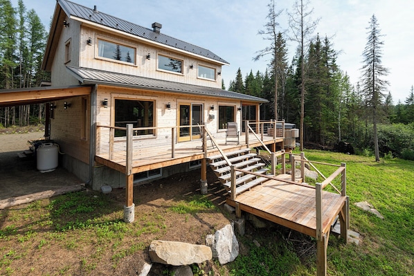 A two-story wooden cabin with a deck, a staircase, and a view of the surrounding forest.