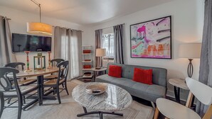 A modern living room with a grey sofa, a round coffee table, a dining table with chairs, a TV, and a large abstract painting on the wall.