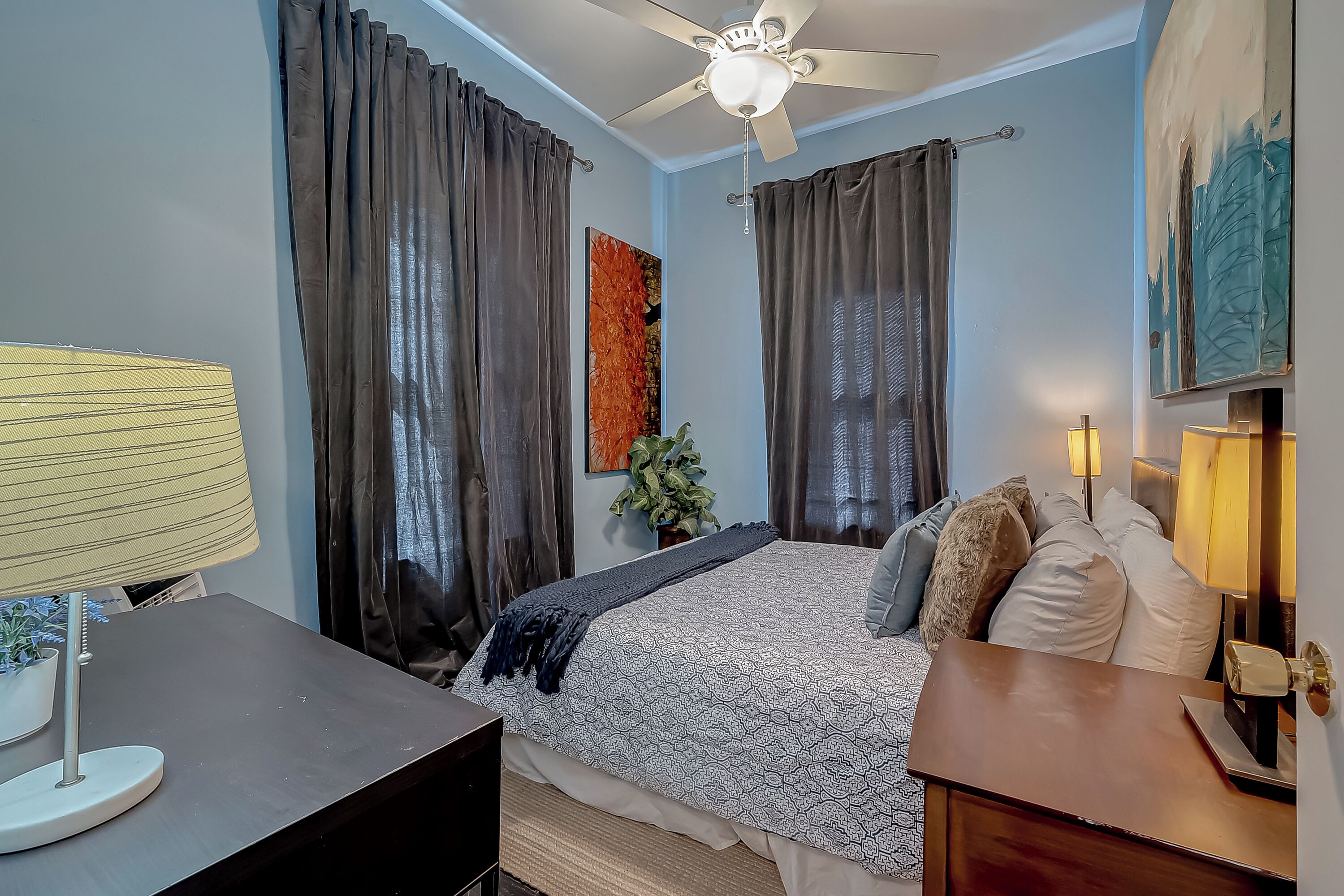 Une chambre à coucher avec un lit, des rideaux, un ventilateur de plafond, un bureau et une lampe.