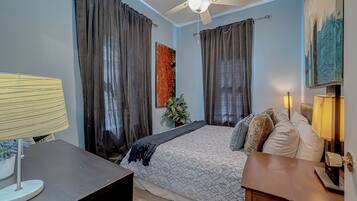Une chambre à coucher avec un lit, des rideaux, un ventilateur de plafond, un bureau et une lampe.