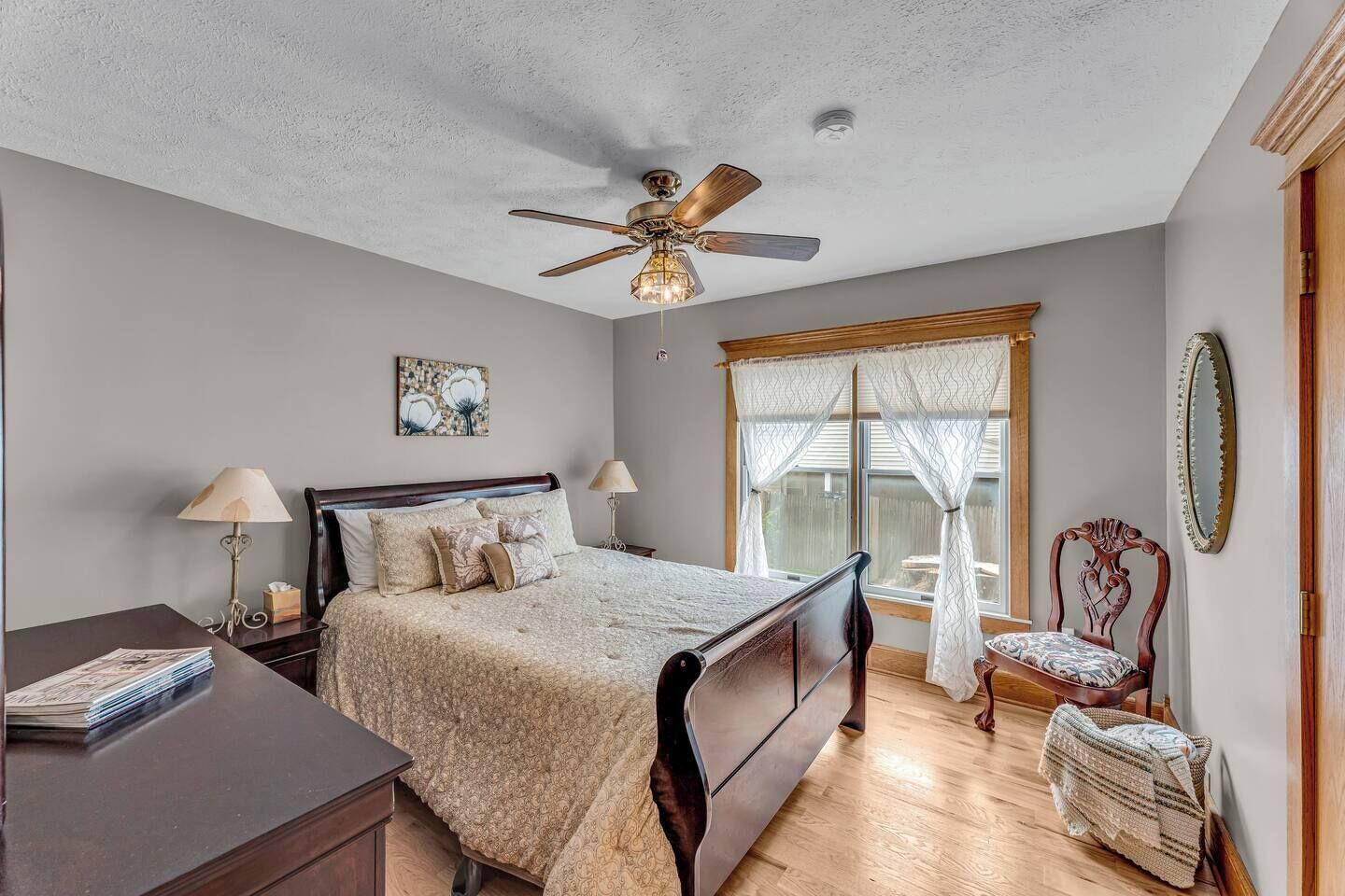 Une chambre à coucher comprenant un lit, des tables de chevet, un bureau, une chaise, un ventilateur de plafond, une fenêtre avec des rideaux et un miroir.