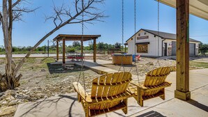 A wooden swing set with two chairs, a covered seating area, and a building in the background. - Hot Tub + Fire Pit: Possum Kingdom Lake Retreat! (Strawn)