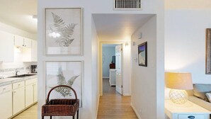A hallway with framed botanical prints, a wicker chair, a white cabinet, and a lamp on a bedside table. - Beach Access Across the Street, Shared Heated Pool (Corpus Christi)