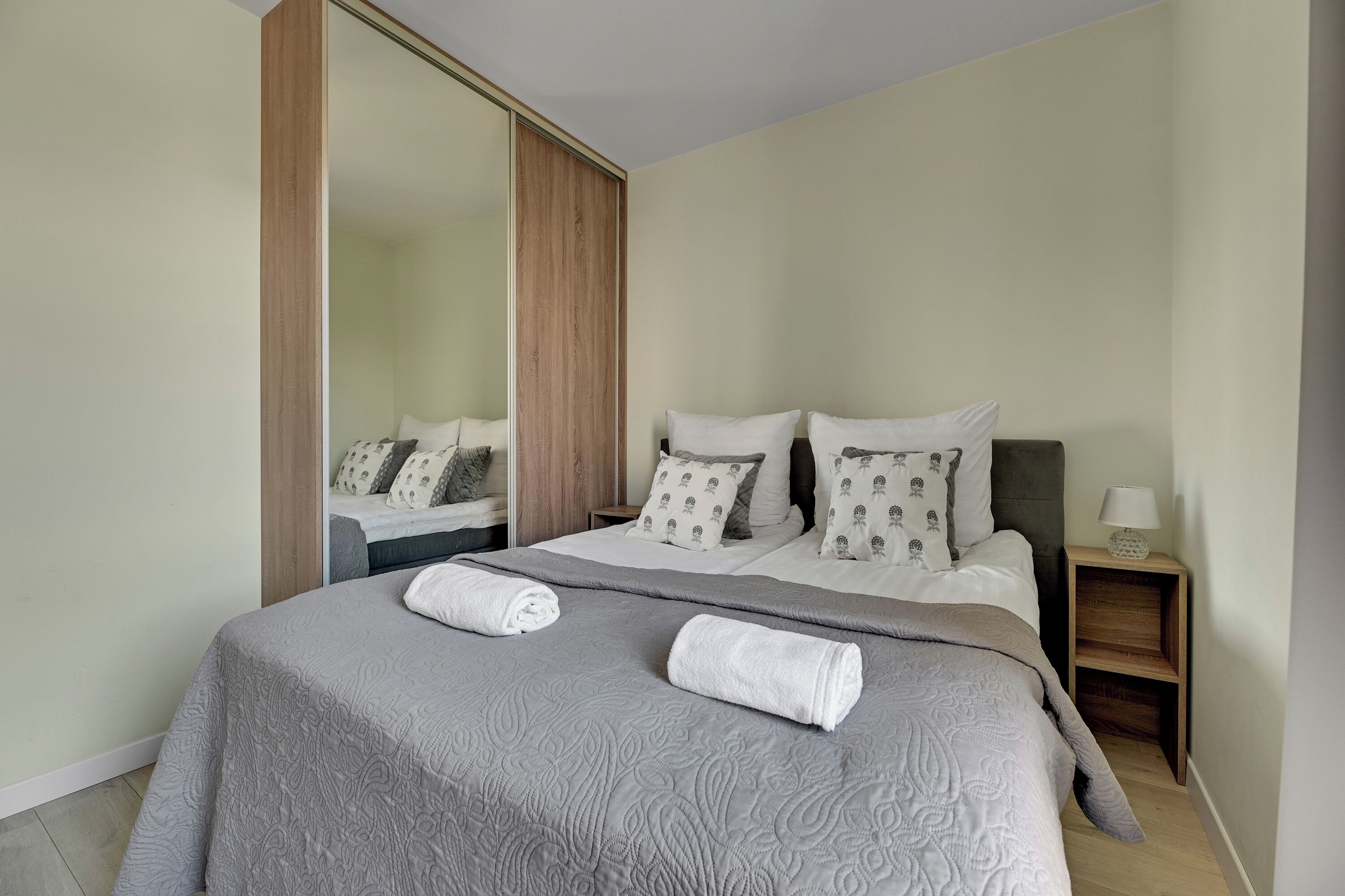Ein ordentlich gemachtes Bett mit einer grauen, gemusterten Tagesdecke und zwei aufgerollten Handtüchern. Am hölzernen Nachttisch stehen eine Lampe und ein Spiegel.
