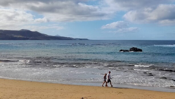 A beach with a rocky outcrop in the water, a clear sky, and a distant mountain range.