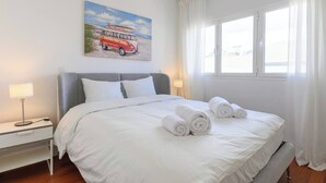 A neatly made bed with white linens and rolled towels, a bedside table with a lamp, a painting of a vintage car on the wall, and a window with curtains.