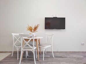 Un coin repas avec une table ronde, deux chaises blanches, un vase contenant des plantes séchées et un téléviseur à écran plat fixé au mur.