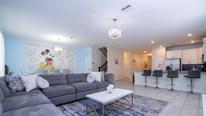 A modern living room with a gray sectional sofa, a white coffee table, and a blue patterned rug. There is a kitchen area with white cabinets and a stainless steel refrigerator. - Spacious Themed 8BR Villa at Champions Gate – Resort full Access, Near Disney! (Four Corners)