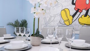A dining table set with white plates, bowls, and wine glasses, accompanied by a potted plant and a Mickey Mouse poster on the wall. - Spacious Themed 8BR Villa at Champions Gate – Resort full Access, Near Disney! (Four Corners)
