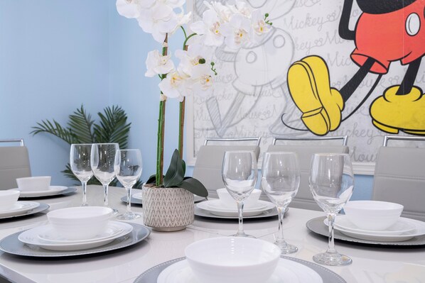 A dining table set with white plates, bowls, and wine glasses, accompanied by a potted plant and a Mickey Mouse poster on the wall. - Spacious Themed 8BR Villa at Champions Gate – Resort full Access, Near Disney! (Four Corners)
