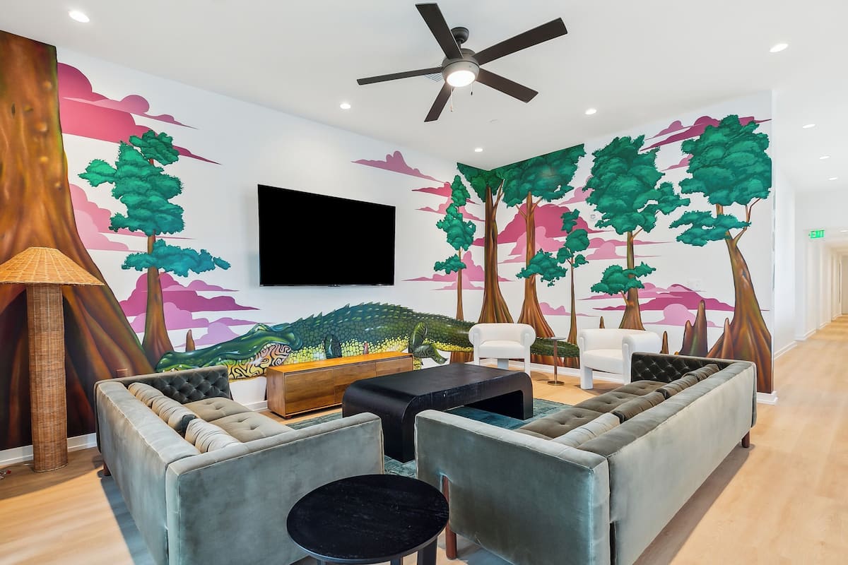 A modern living room with a large flat-screen TV mounted on the wall, a ceiling fan, and a mural of trees and mountains. There are plush sofas and a coffee table, with a lamp and a sideboard in the corner.