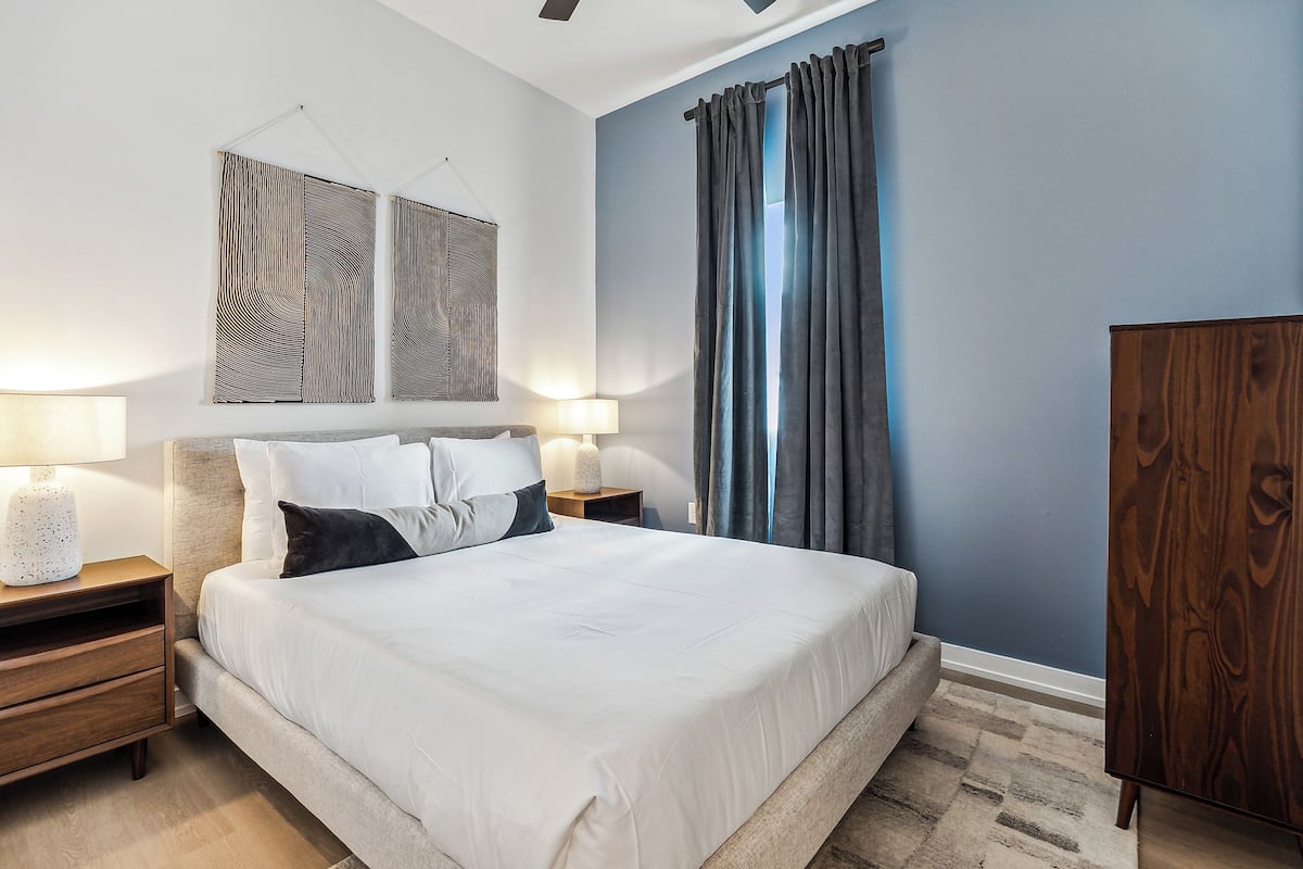 A bedroom with a bed, two bedside tables with lamps, a wooden nightstand, a wardrobe, and a large window with curtains.