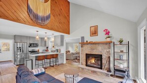 A modern kitchen with stainless steel appliances, a central island with bar stools, a stone fireplace, and a living area with a blue sofa and a round coffee table.