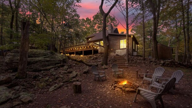A cabin with a deck, surrounded by trees, and a fire pit with chairs.