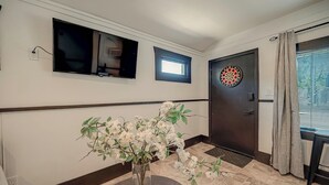 Un couloir avec un téléviseur, une petite fenêtre, une porte sur laquelle est accroché un jeu de fléchettes et un vase rempli de fleurs.