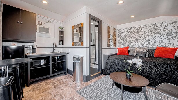 Un espace de vie compact comprenant un coin cuisine, un canapé avec des coussins rouges et un mur décoré d’un motif.