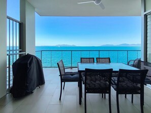 Een balkon met een tafel, stoelen, een barbecue en uitzicht op de zee en verre bergen.