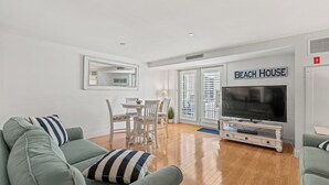 A living room with a large flat-screen TV, a dining area with a table and chairs, and a sign reading "BEACH HOUSE".