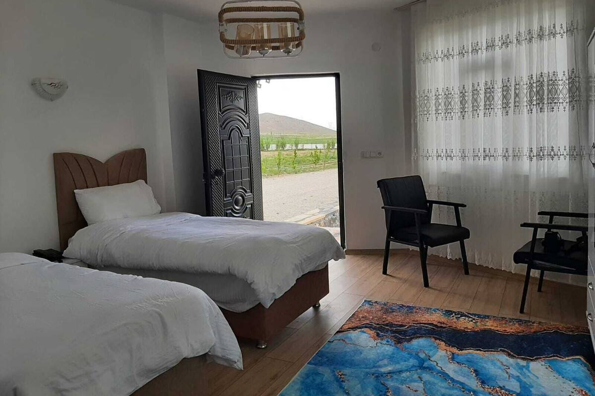 A bedroom with two beds, a chair, a desk, and a view of the outdoors.