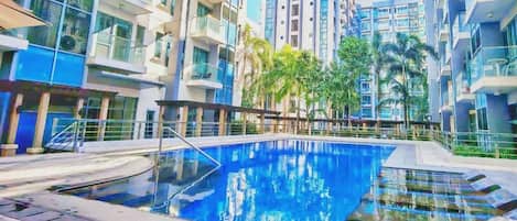 Une piscine entourée de bâtiments modernes dotés de balcons, et un panneau indiquant que l’accès à la piscine est gratuit pour trois personnes.