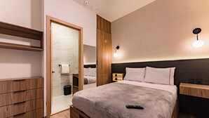Una habitación de hotel moderna con una cama grande, muebles de madera, un armario empotrado y un baño con mampara de cristal.