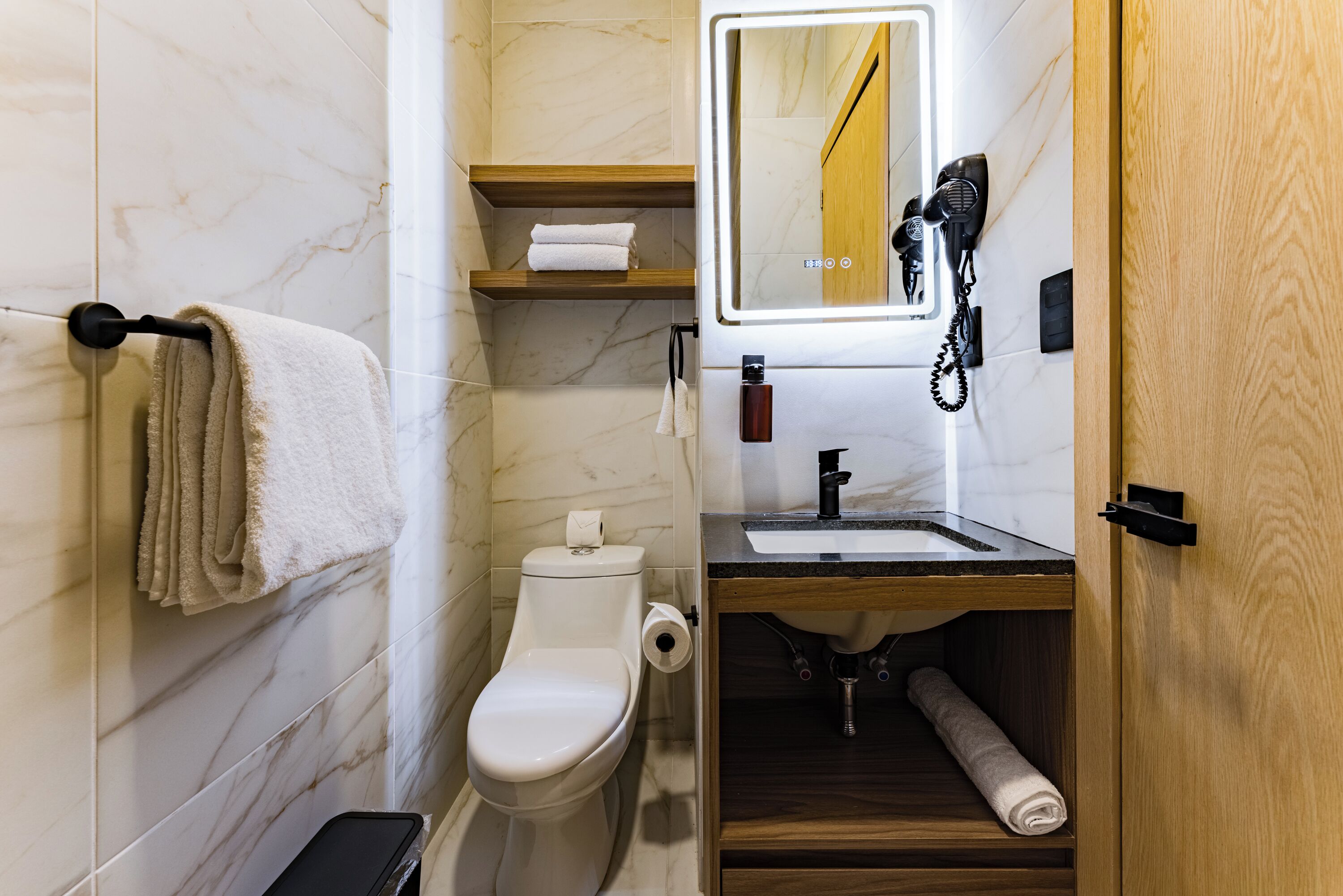 A modern bathroom with a toilet, sink, and mirror. There are towels on a rack, a hairdryer, and a bottle on the counter.