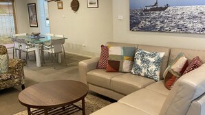 Un salon avec un canapé beige, une table à manger en verre et des chaises. Un tableau représentant un navire est accroché au mur.