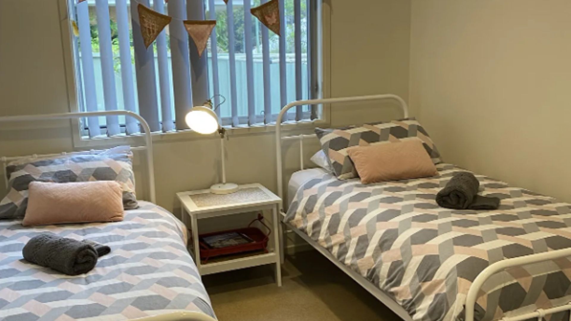 A bedroom with two beds, each with patterned bedding and pillows. There is a bedside table with a lamp and a red basket.