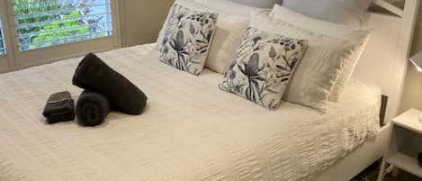 A neatly made bed with white bedding and floral-patterned pillows, a small bedside table, and a window with a view of greenery.